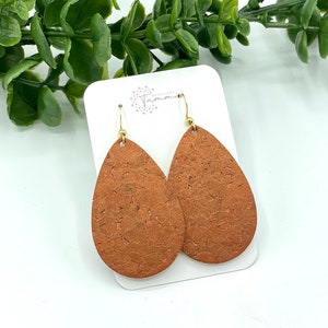 May include: A pair of teardrop-shaped earrings made of cork with a brown, speckled pattern. The earrings have gold-colored hooks.