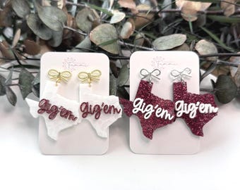 May include: Two pairs of Texas-shaped earrings. One pair is white with the words "Gig'em" in white script. The other pair is maroon glitter with the words "Gig'em" in white script. Both pairs have bow-shaped earring hooks.