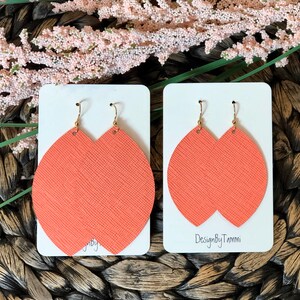 May include: A pair of coral-colored leather earrings with gold hooks. The earrings are shaped like a teardrop and have a textured surface. The earrings are displayed on a white card with the text "DesignByTammi" printed on it.