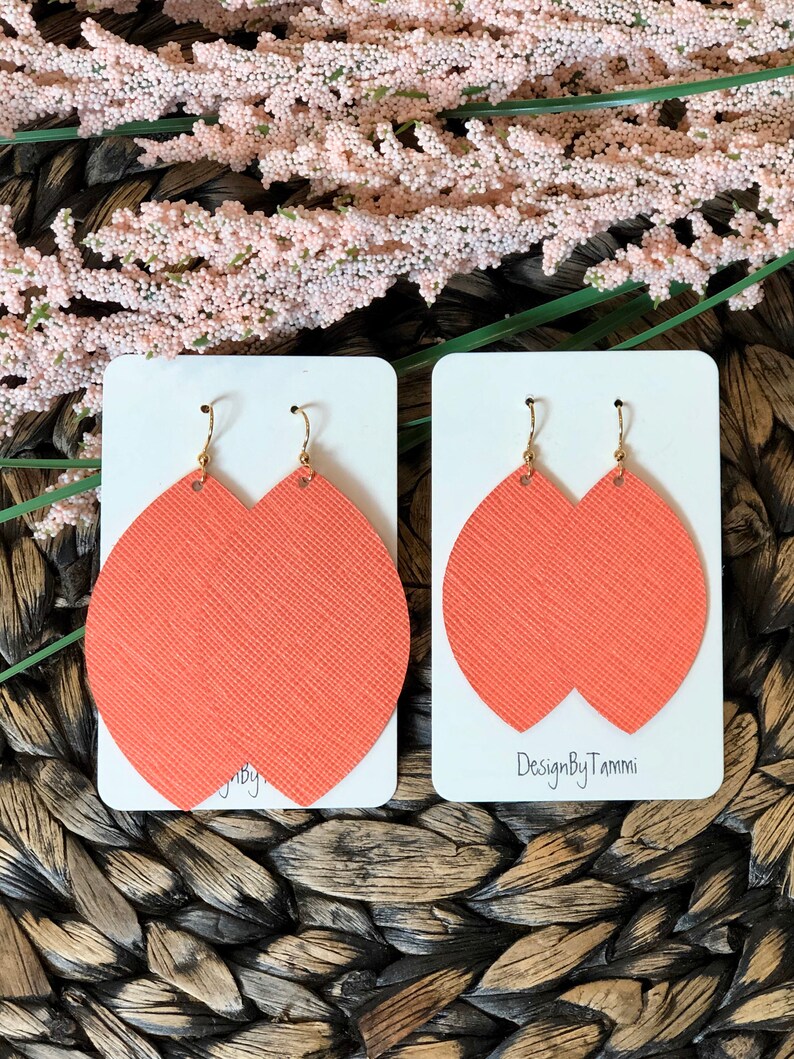 May include: A pair of coral-colored leather earrings with gold hooks. The earrings are shaped like a teardrop and have a textured surface. The earrings are displayed on a white card with the text "DesignByTammi" printed on it.