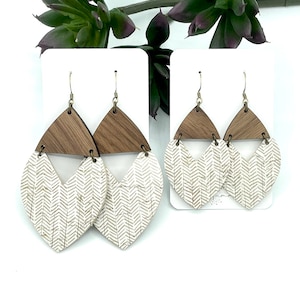 May include: A pair of wooden and cork earrings with a chevron pattern. The earrings are a light brown color with a white chevron pattern. The earrings are hanging from gold hooks.