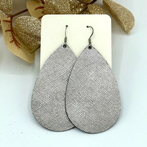 May include: A pair of silver teardrop-shaped earrings with a textured finish. The earrings are made of leather and have silver hooks.