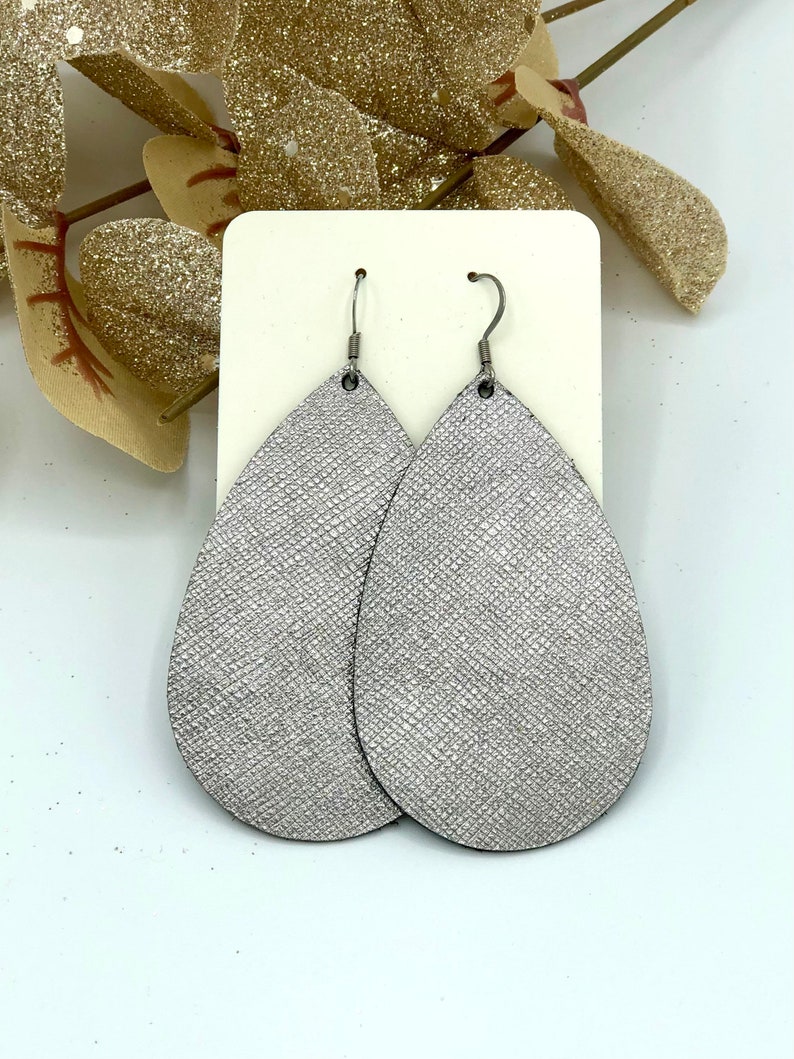 May include: A pair of silver teardrop-shaped earrings with a textured finish. The earrings are made of leather and have silver hooks.