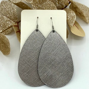 May include: A pair of silver teardrop-shaped earrings with a textured finish. The earrings are made of leather and have silver hooks.