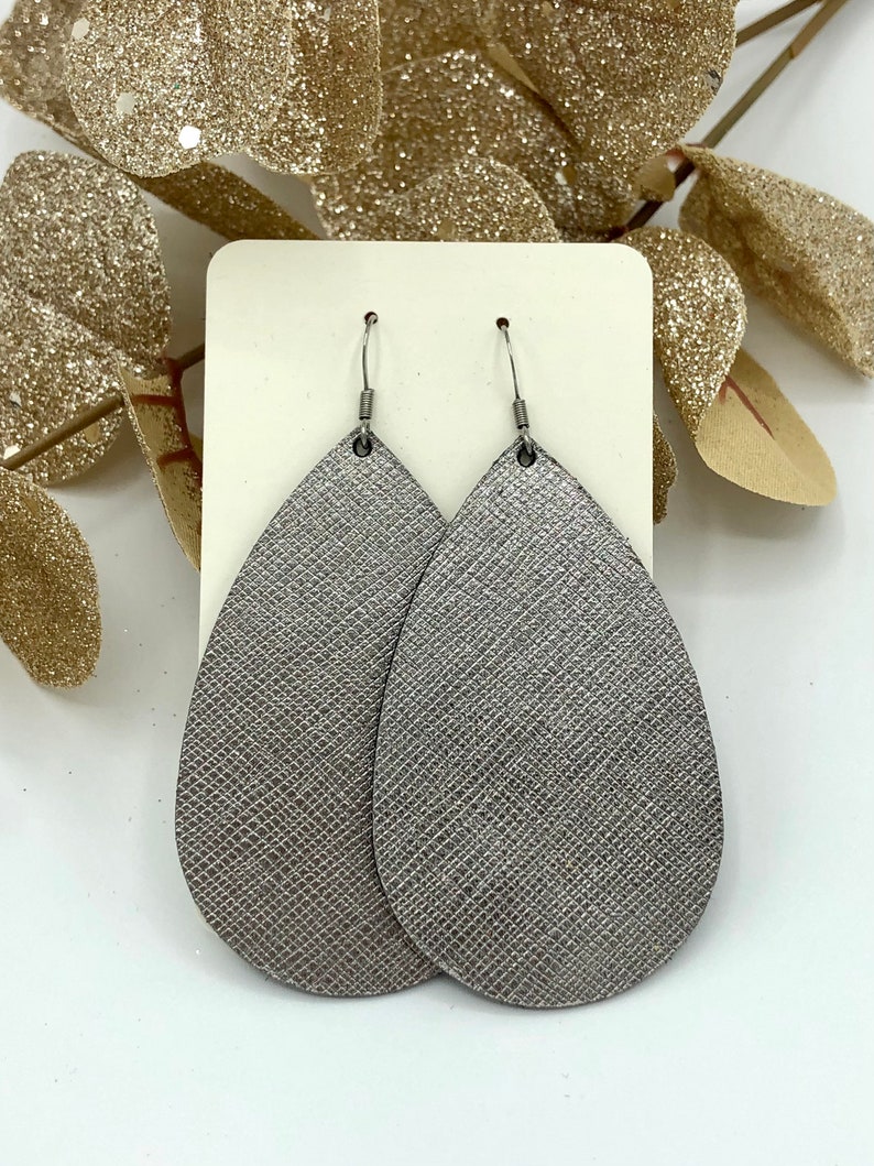 May include: A pair of silver teardrop-shaped earrings with a textured finish. The earrings are made of leather and have silver hooks.