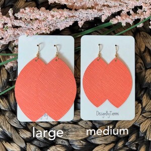 May include: Two pairs of coral-colored leather earrings, one pair labeled "large" and the other labeled "medium". The earrings are shaped like a teardrop with a rounded top and a pointed bottom. The earrings are on white cards with the text "DesignByTammi" on the medium card.
