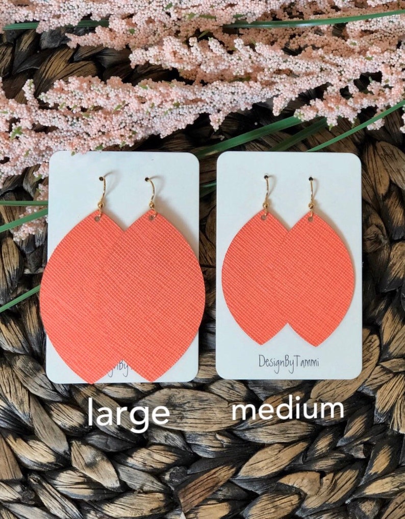 May include: Two pairs of coral-colored leather earrings, one pair labeled "large" and the other labeled "medium". The earrings are shaped like a teardrop with a rounded top and a pointed bottom. The earrings are on white cards with the text "DesignByTammi" on the medium card.