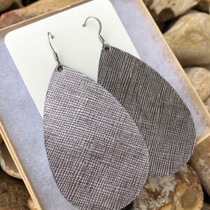 May include: A pair of silver teardrop-shaped earrings with a textured surface. The earrings are made of leather and have silver hooks.