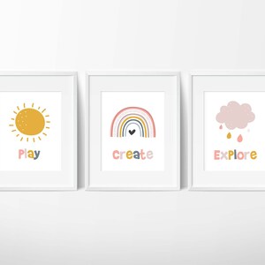 May include: Three white framed prints with colorful illustrations and text. The first print features a yellow sun with the word "Play" below it. The second print features a rainbow with a heart at the bottom and the word "Create" below it. The third print features a pink cloud with raindrops and the word "Explore" below it.