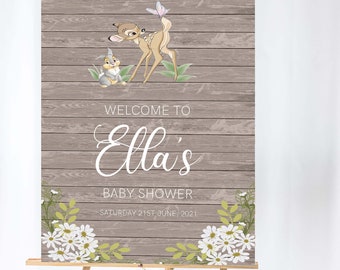 May include: A wooden sign on an easel, featuring a baby shower announcement. The sign has a rustic wood grain background with illustrations of a deer and a rabbit. Text reads "Welcome to Ella's Baby Shower" with the date "Saturday 21st June, 2021".