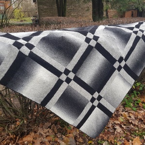 Puede incluir: Una manta de punto negra y blanca con un patrón geométrico de cuadrados y rombos. La manta está drapeada sobre un arbusto con hojas marrones.