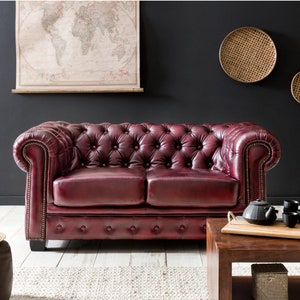 Op de afbeelding: Een dieprode leren chesterfieldbank met knoopvormige bekleding en gerolde armleuningen. De bank staat in een woonkamer met een houten salontafel en decoratieve wanddecoraties.