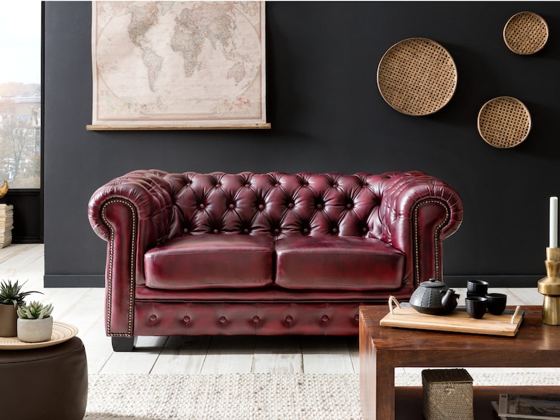 Op de afbeelding: Een dieprode leren chesterfieldbank met knoopvormige bekleding en gerolde armleuningen. De bank staat in een woonkamer met een houten salontafel en decoratieve wanddecoraties.