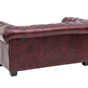Op de afbeelding: Een bordeauxrode leren bank met gestoffeerde bekleding en een gerolde rugleuning. De bank heeft een klassiek ontwerp en is perfect voor een traditionele woonkamer.