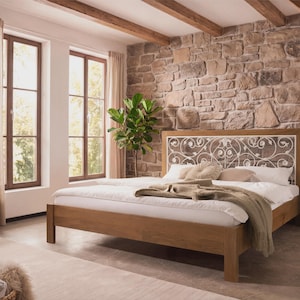 Könnte beinhalten: Ein Holzbettrahmen mit einem weißen Kopfteil mit einem dekorativen, wirbelnden Metalldesign. Das Bett ist mit weißer Bettwäsche und Kissen sowie einer beigefarbenen Decke bezogen. Der Raum hat eine Steinmauer und große Fenster.