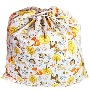 May include: A white drawstring bag with a woodland animal print. The print features foxes, deer, owls, rabbits, and hedgehogs in orange, brown, and yellow against a white background. The bag is made of cotton and has a drawstring closure.
