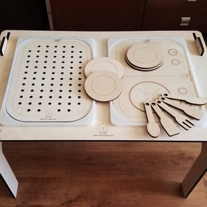 Può includere: Un set da cucina in legno per bambini con un lavandino, un piano cottura e utensili. Il set è realizzato in legno chiaro e ha una superficie bianca. Il lavandino ha uno scarico e il piano cottura ha due bruciatori. Gli utensili includono un cucchiaio, una spatola e una forchetta.