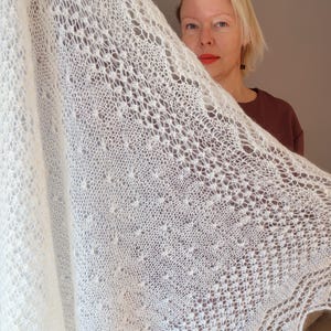 Może przedstawiać: Kremowy szal o otwartym splocie z delikatnym, koronkowym wzorem. Szal jest trzymany w górze, prezentując swój skomplikowany design i fakturę. Dzianina lub szydełkowa tkanina ma miękki, przewiewny wygląd, idealny do warstw.