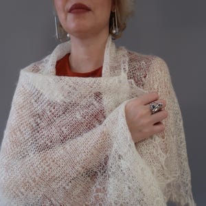 Puede incluir: Un chal de encaje blanco con un patrón delicado. El chal está drapeado sobre los hombros de una persona y está hecho de una tela suave y ligera.