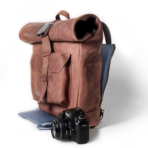 Könnte beinhalten: Ein brauner Leder-Rolltop-Rucksack mit einer großen Fronttasche und einem blauen Futter. Vor dem Rucksack liegt eine schwarze Kamera.