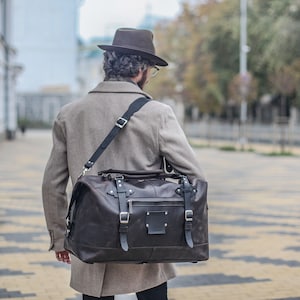Puede incluir: Una persona con un sombrero fedora marr&oacute;n y un abrigo beige lleva una bolsa de viaje de cuero marr&oacute;n con una correa negra.