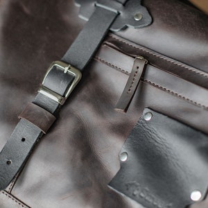 Puede incluir: Primer plano de un bolso de mensajero de cuero marr&oacute;n con una correa de cuero negro y una hebilla plateada. El bolso tiene un bolsillo con cremallera y un peque&ntilde;o bolsillo de parche con una etiqueta cosida.