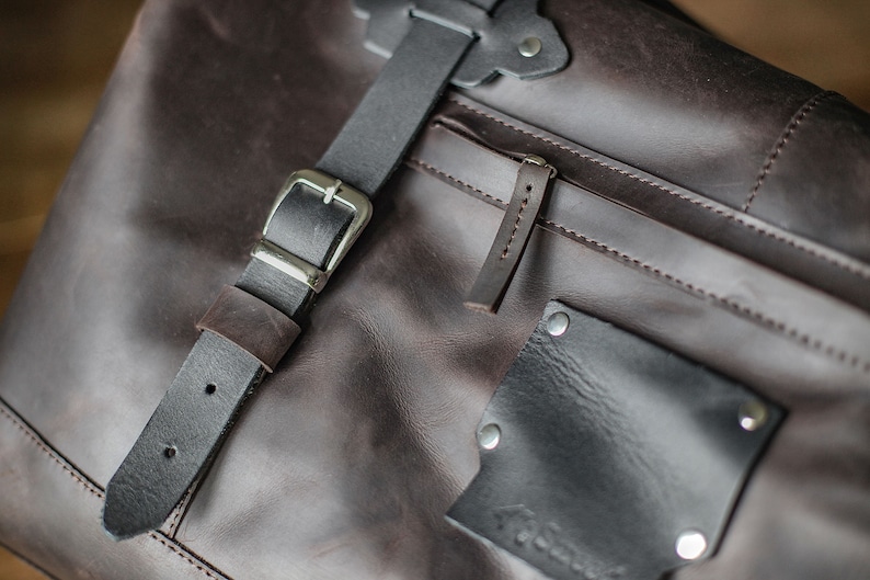 Puede incluir: Primer plano de un bolso de mensajero de cuero marr&oacute;n con una correa de cuero negro y una hebilla plateada. El bolso tiene un bolsillo con cremallera y un peque&ntilde;o bolsillo de parche con una etiqueta cosida.