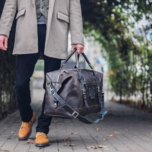 Puede incluir: Una bolsa de viaje de cuero marr&oacute;n con una correa larga y hebillas plateadas. La bolsa la lleva una persona que lleva un abrigo gris y pantalones oscuros.