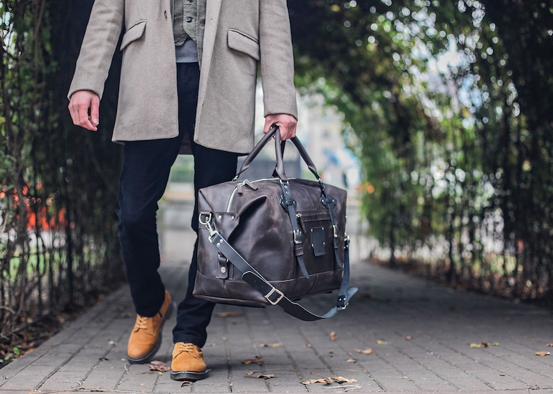 Puede incluir: Una bolsa de viaje de cuero marr&oacute;n con una correa larga y hebillas plateadas. La bolsa la lleva una persona que lleva un abrigo gris y pantalones oscuros.