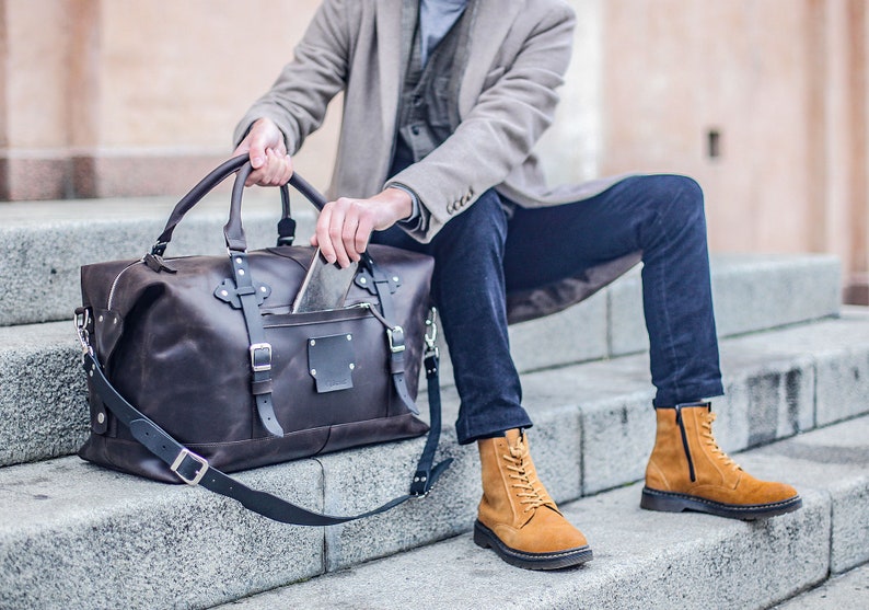 Puede incluir: Una bolsa de viaje de cuero marr&oacute;n con correas y hebillas negras. La bolsa est&aacute; sentada en un conjunto de escalones de concreto. Una persona est&aacute; metiendo la mano en la bolsa.