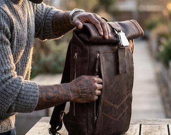 Könnte beinhalten: Ein brauner Lederrucksack mit Rollverschluss und silberner Schnalle. Der Rucksack hat mehrere Reißverschlusstaschen und verstellbare Träger. Die Tasche ist aus einem Leder mit Used-Look gefertigt und liegt auf einer Holzoberfläche.