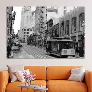 May include: Black and white print of a San Francisco street scene featuring a cable car on tracks. Buildings line the street, and the image is displayed above an orange sofa with pillows and small side tables.