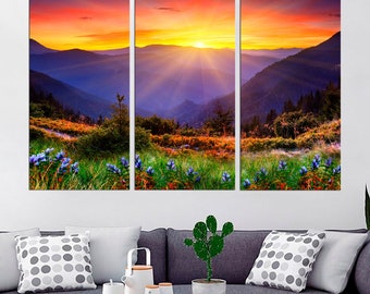 May include: A three-panel canvas print depicting a colorful sunset over a mountain range. The sun is setting behind the mountains, casting a warm glow over the landscape. The mountains are covered in lush green trees and wildflowers.