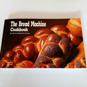 May include: A cookbook titled "The Bread Machine Cookbook" by Donna Rathmell German. The cover features a close-up of various types of baked bread, with a focus on golden-brown loaves and rolls. The book is on a white surface.