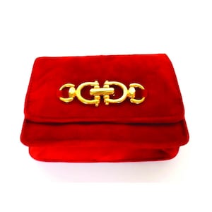 Op de afbeelding: Een rode fluwelen clutch met een goudkleurige sluiting. De rechthoekige tas heeft een overslagklep en een decoratieve gouden sluiting in het midden. De tas staat tegen een witte achtergrond.