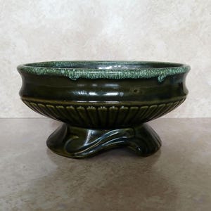 May include: A dark olive green ceramic bowl with a pedestal base. The bowl features a wide, fluted body with vertical ridges and a distinct, mottled light green drip glaze along the upper rim. The base has a curved, sculptural design.