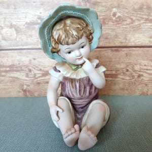 May include: A porcelain figurine of a seated child wearing a lavender dress and a light blue bonnet. The child has blonde hair and is touching their index finger to their lips. The figurine has a vintage aesthetic and is set against a wooden background.
