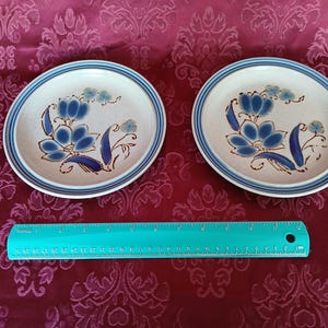 K&ouml;nnte beinhalten: Zwei Keramikplatten mit floralem Design. Die Platten haben eine cremefarbene Basis mit blauen Blumenmustern und einen blau-braunen Rand. Ein t&uuml;rkisfarbenes Lineal ist darunter platziert.