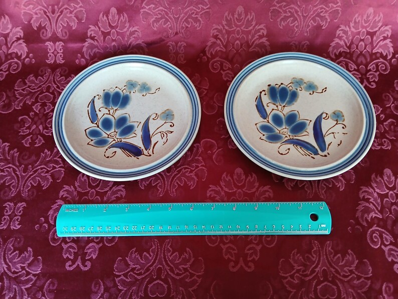 K&ouml;nnte beinhalten: Zwei Keramikplatten mit floralem Design. Die Platten haben eine cremefarbene Basis mit blauen Blumenmustern und einen blau-braunen Rand. Ein t&uuml;rkisfarbenes Lineal ist darunter platziert.