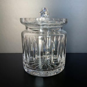 May include: A clear glass jar with a matching lid featuring a faceted knob handle. The cylindrical body is decorated with vertical oval cut patterns and a starburst design on the base, showcasing a reflective and transparent crystalline texture.