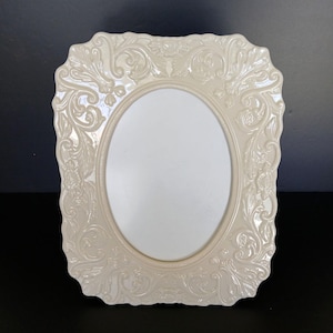 May include: A cream-colored rectangular picture frame with an oval center opening. The frame features a glossy finish and intricate, raised scrollwork and floral relief patterns covering the entire border surrounding the central oval area.