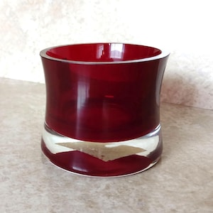 May include: A cylindrical glass candle holder featuring a deep ruby red upper section and a thick, clear glass base. The vessel has smooth, curved walls and a wide, open top, showcasing a vibrant contrast between the transparent red and clear glass.
