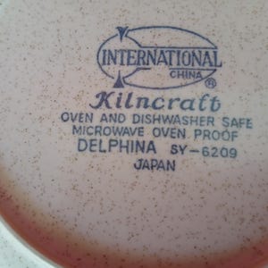 K&ouml;nnte beinhalten: Nahaufnahme eines beigefarbenen Kilncraft Delphina-Tellers mit einem gesprenkelten Muster. Der Teller hat einen rosa Rand und tr&auml;gt die Aufschrift "INTERNATIONAL CHINA", "Kilncraft", "OVEN AND DISHWASHER SAFE", "MICROWAVE OVEN PROOF", "DELPHINA SY-6209" und "JAPAN".