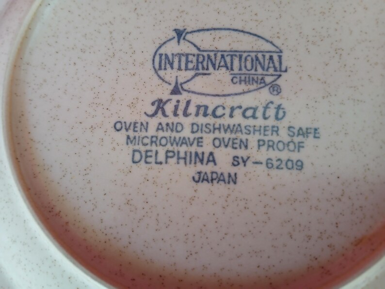 K&ouml;nnte beinhalten: Nahaufnahme eines beigefarbenen Kilncraft Delphina-Tellers mit einem gesprenkelten Muster. Der Teller hat einen rosa Rand und tr&auml;gt die Aufschrift "INTERNATIONAL CHINA", "Kilncraft", "OVEN AND DISHWASHER SAFE", "MICROWAVE OVEN PROOF", "DELPHINA SY-6209" und "JAPAN".