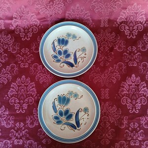 K&ouml;nnte beinhalten: Zwei Keramikplatten mit floralem Design. Die Teller haben eine cremefarbene Basis mit blauen Blumenmustern und einem blauen Rand. Die Teller liegen auf einer roten, gemusterten Oberfl&auml;che.