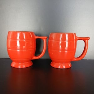 May include: Two bright orange ceramic mugs with a barrel-like shape, featuring horizontal embossed lines around the body, a sturdy side handle, and a pedestal base. The mugs have a smooth, glossy finish and a consistent, vibrant orange color.