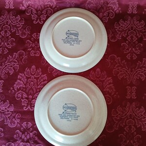 Puede incluir: Dos platos de cer&aacute;mica blanquecina con borde beige. Los platos tienen el texto "International" y "Kilncraft" impreso. Los platos est&aacute;n sobre una tela burdeos estampada.