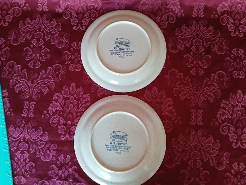 Puede incluir: Dos platos de cer&aacute;mica blanquecina con borde beige. Los platos tienen el texto "International" y "Kilncraft" impreso. Los platos est&aacute;n sobre una tela burdeos estampada.