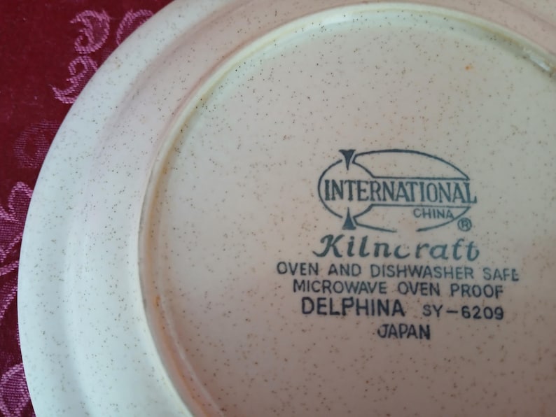 K&ouml;nnte beinhalten: Nahaufnahme eines beigefarbenen Tellers mit gesprenkeltem Muster. Der Teller hat einen breiten Rand und der Text "INTERNATIONAL CHINA Kilncraft OVEN AND DISHWASHER SAFE MICROWAVE OVEN PROOF DELPHINA SY-6209 JAPAN" ist aufgedruckt.
