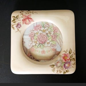 May include: A square ceramic plate with rounded corners and a cream-colored base. The center features a circular illustration of a bouquet of pink and white roses in a vase. Additional floral motifs in pink, purple, and green decorate the top left and bottom right corners of the plate.